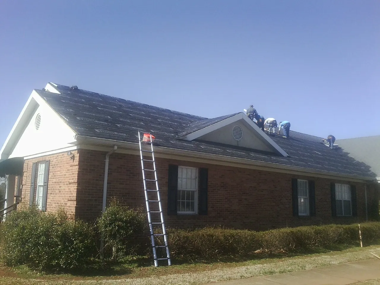 Crew tearing shingles off a brick ranch roof with a ladder set — Metal Roof Repair work by our crew in Manchester